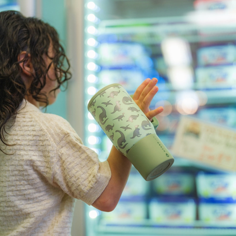 Person holding a green thermos with dinosaur design in front of a refrigerator.