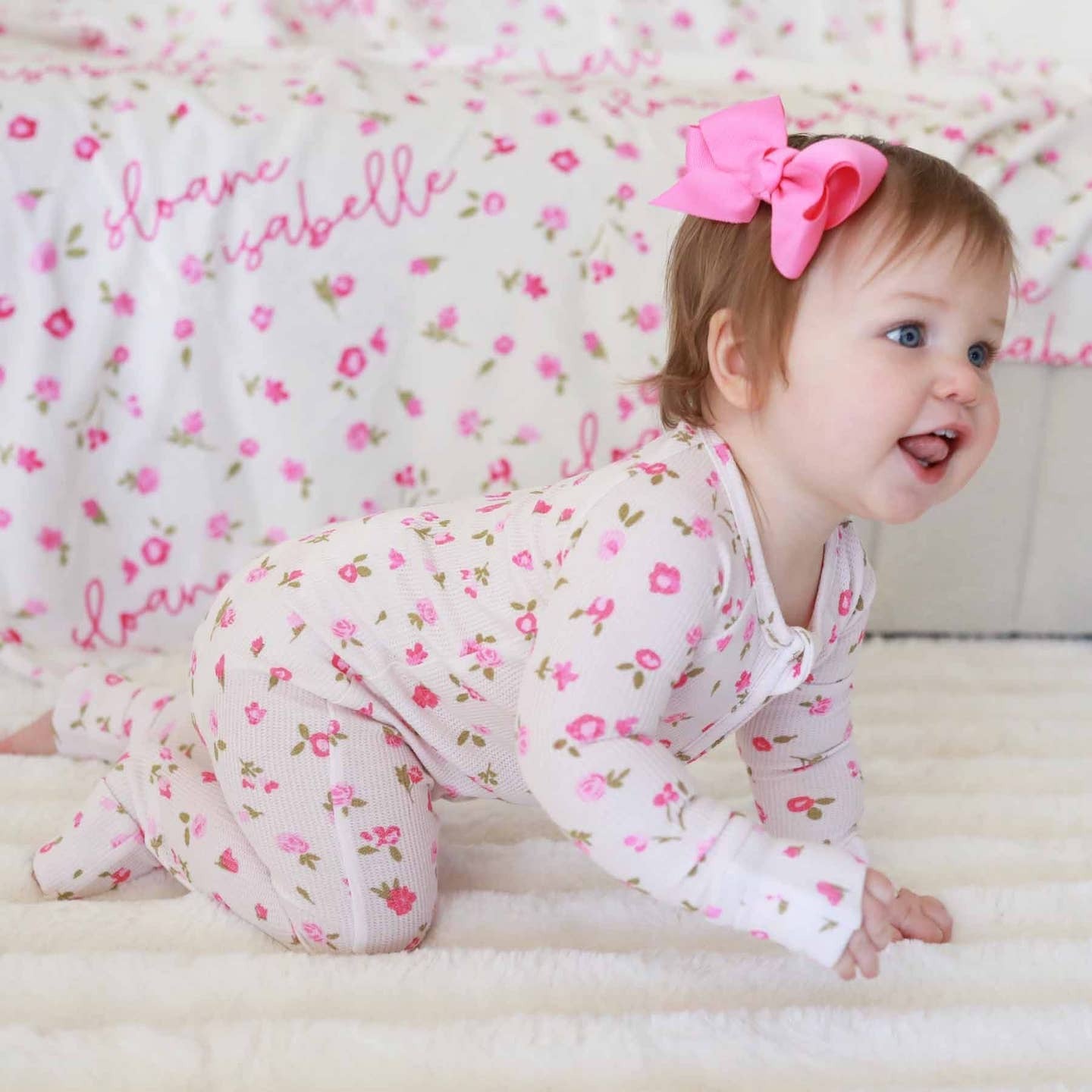 Baby in a floral outfit with a pink bow sitting on a white surface.