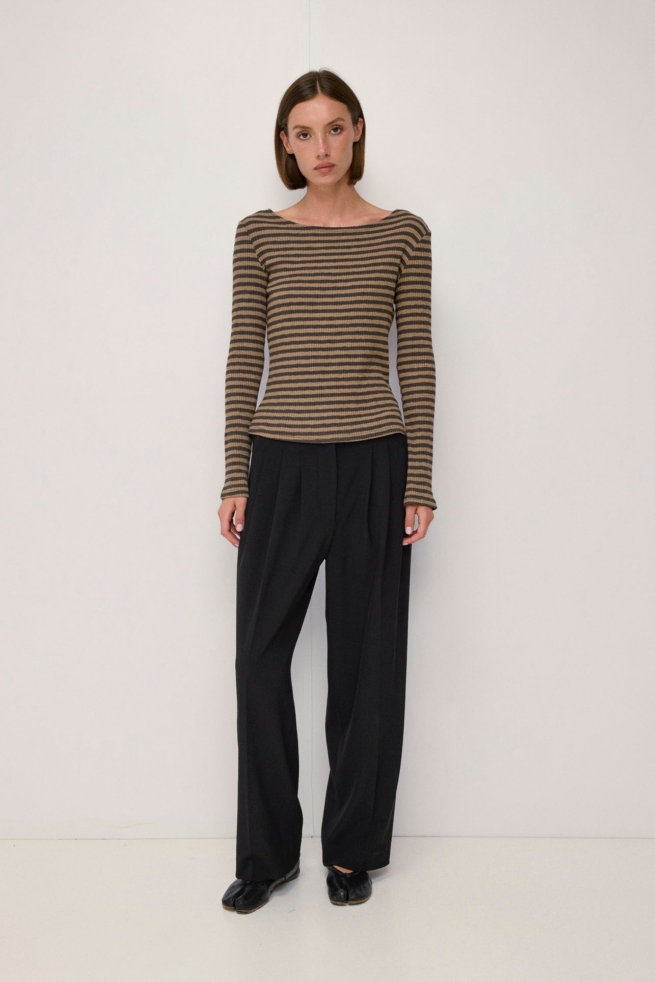 A model stands against a plain white studio background wearing a brown and beige horizontally striped long-sleeve boat-neck top, black wide-leg trousers, and black shoes.