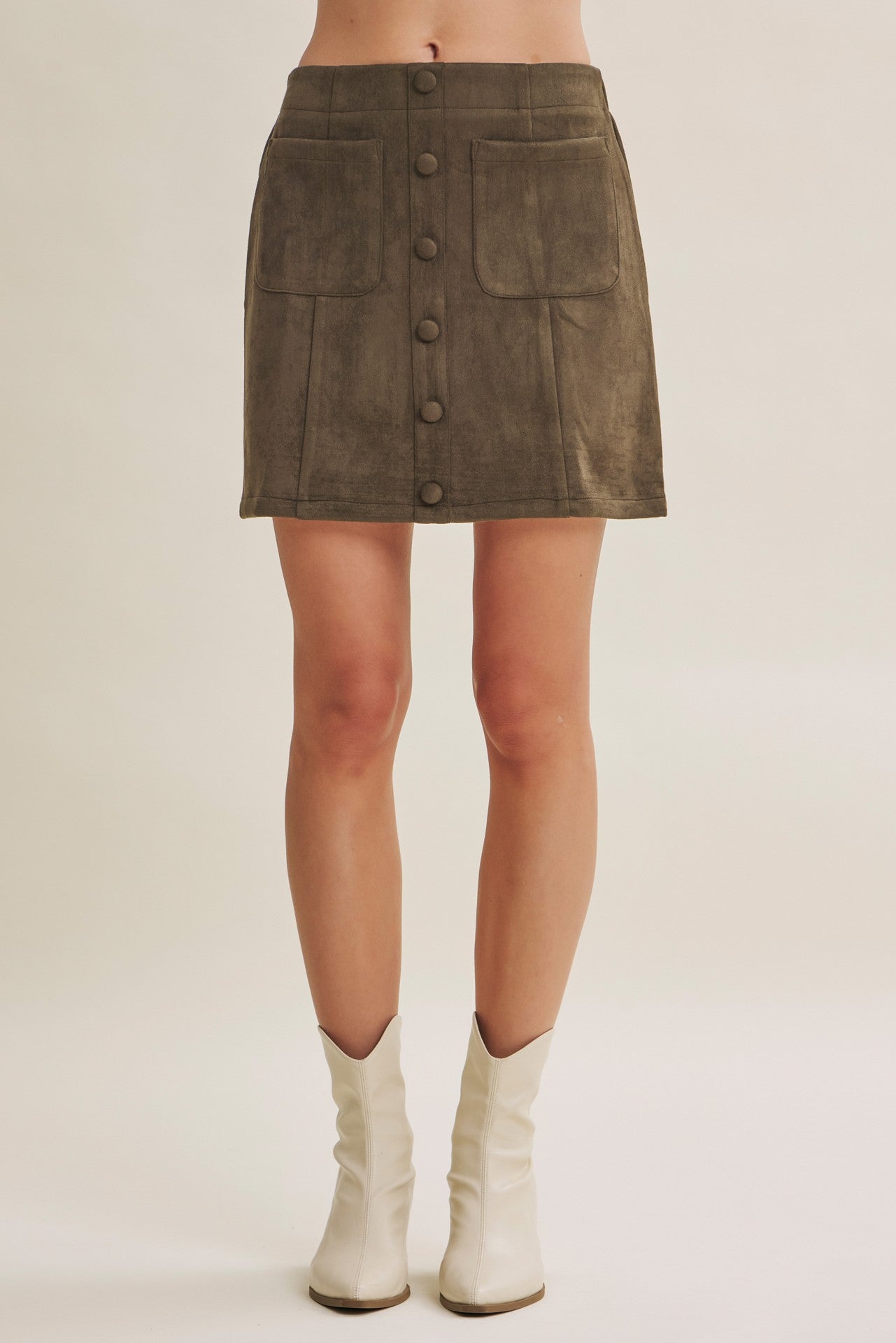A model from the waist down wearing a dark brown suede mini skirt with front button details and flap pockets, paired with off-white cowboy boots, against a light beige studio background.