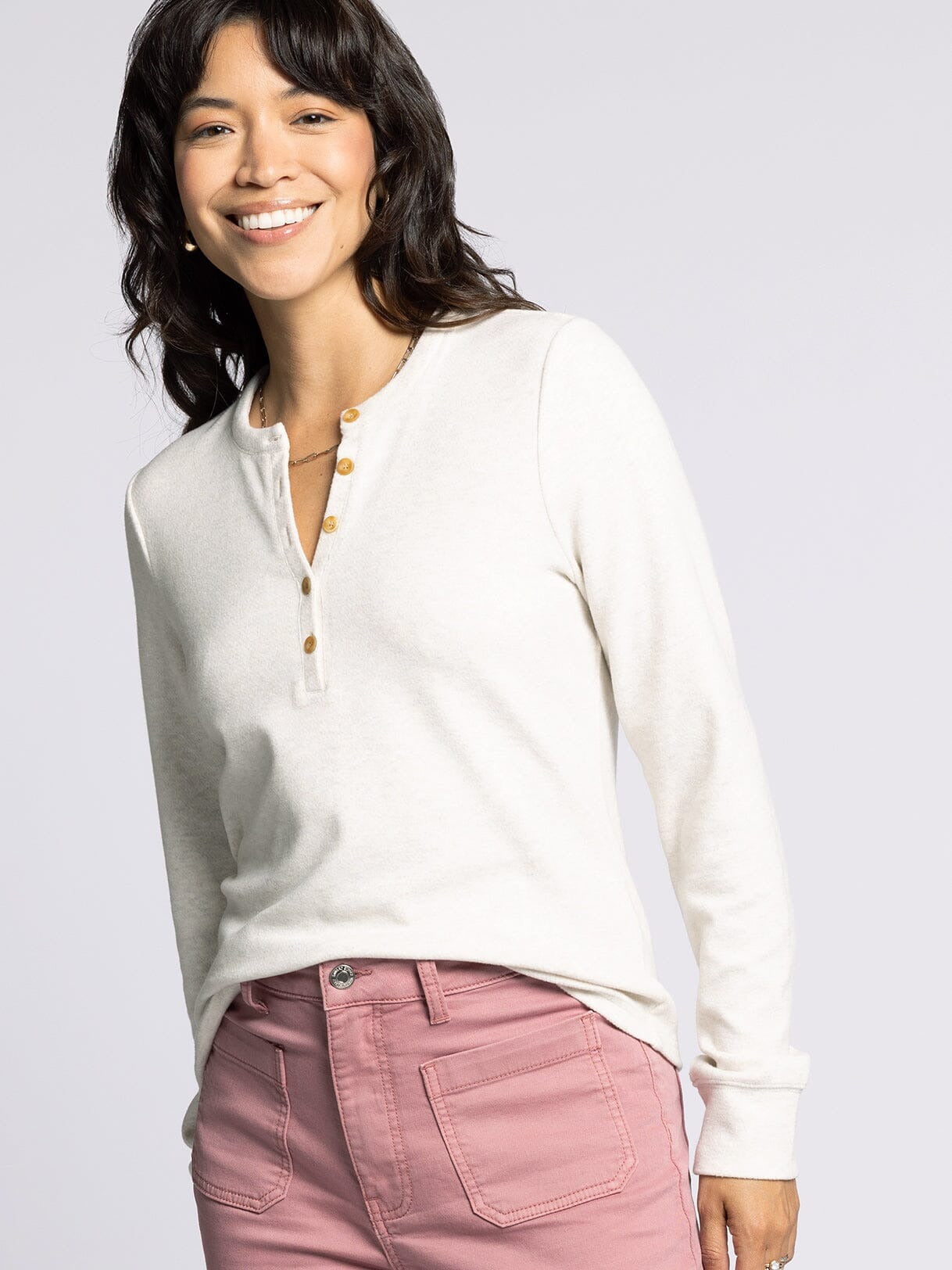 Woman wearing a white long-sleeve shirt with gold buttons and pink pants on a light gray background