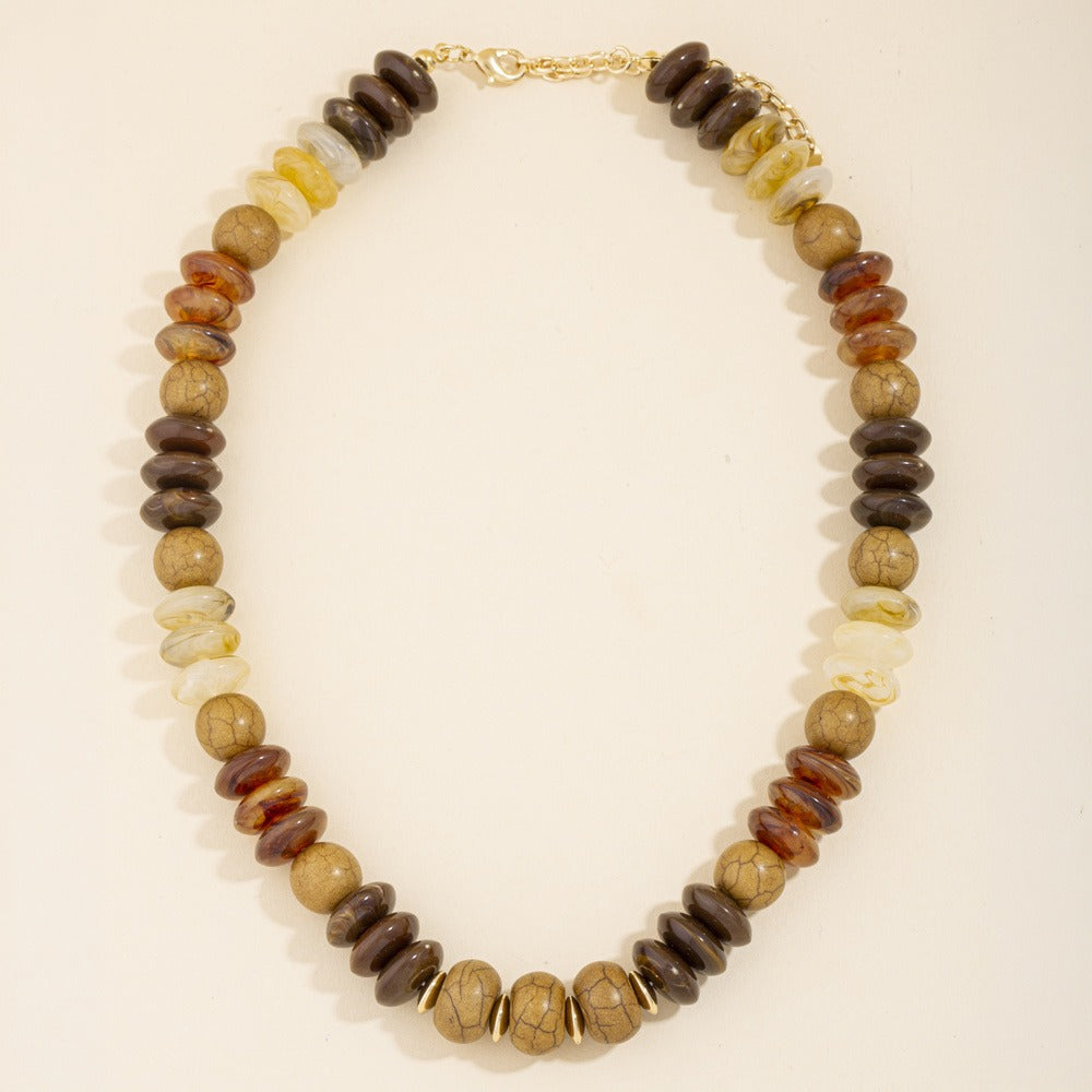 A beaded necklace made of flat, disc-like stone beads in earthy brown, tan, cream, and dark brown tones, with a gold clasp, photographed on a plain light background.