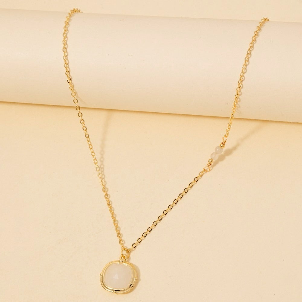 A delicate gold chain necklace with a small round white stone pendant, displayed on a cream-colored background.