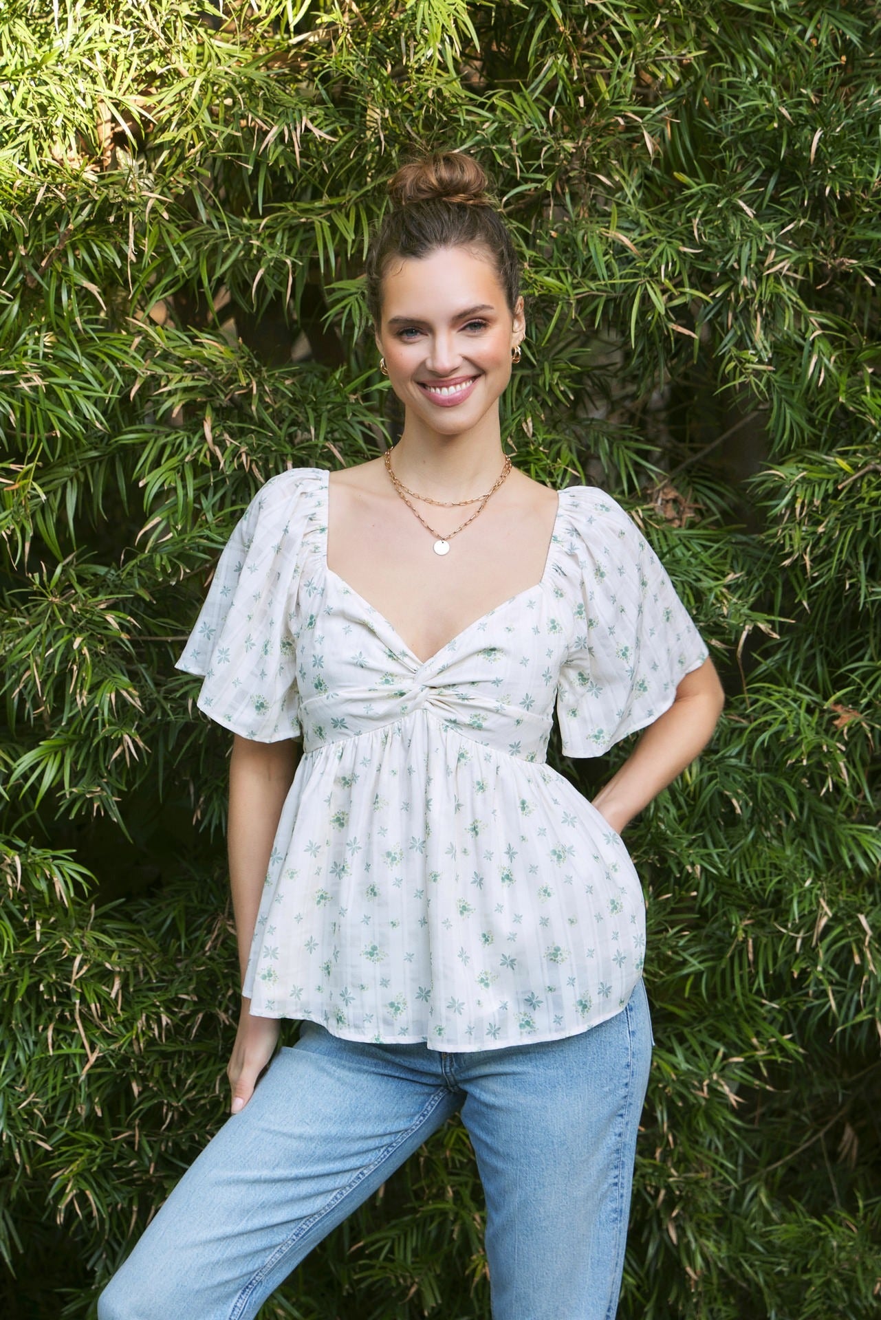 Woman wearing a light green floral blouse and blue jeans standing against a green leafy background