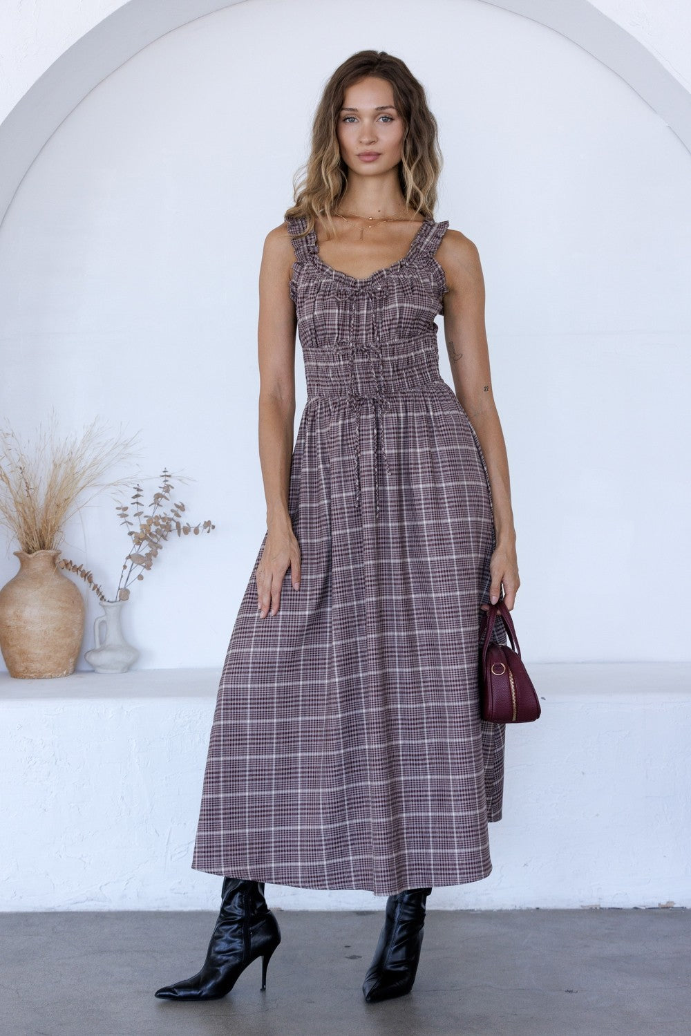 A woman models a sleeveless midi dress with a purple and brown plaid pattern, styled with black pointed-toe heeled boots and a small burgundy handbag, standing in front of a white arched wall.