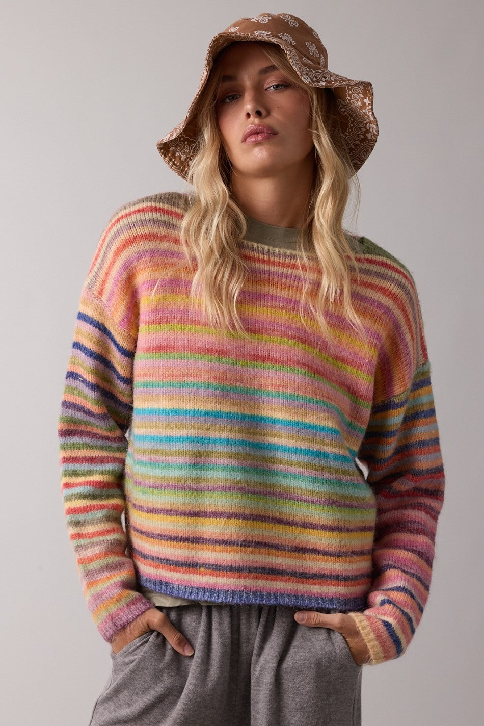 A woman wearing a fuzzy, multicolored striped knit sweater, a brown sequined bucket hat, and gray sweatpants, posing against a neutral light gray studio background.