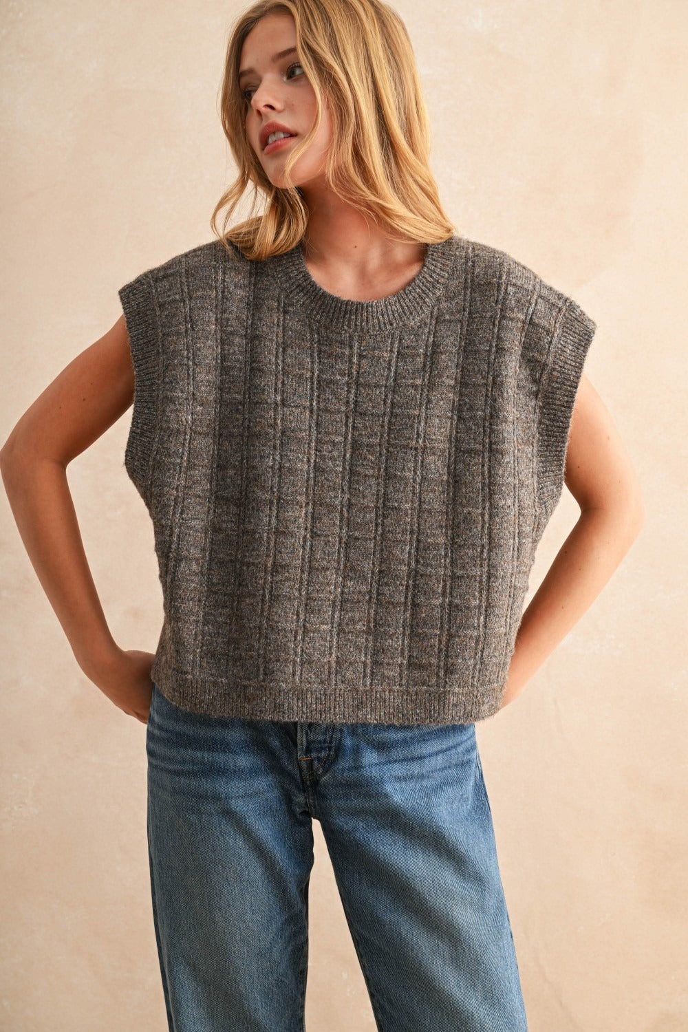 Woman wearing a gray sleeveless knitted top and blue jeans against a beige background