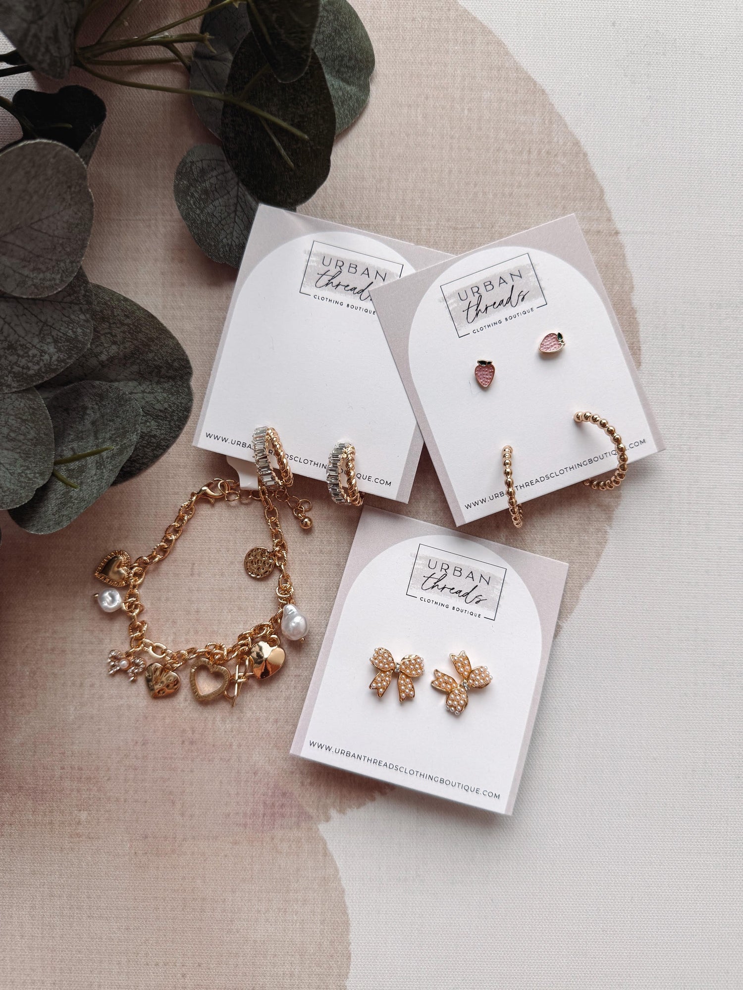 Gold jewelry set including bracelet and earrings on branded cards with a plant in the background.