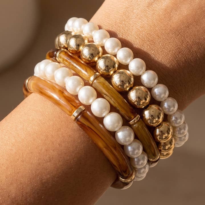 A close-up of a wrist wearing a stack of multi-strand bracelets, including white pearl beads, polished gold-tone metal beads, and translucent amber-brown bangles.