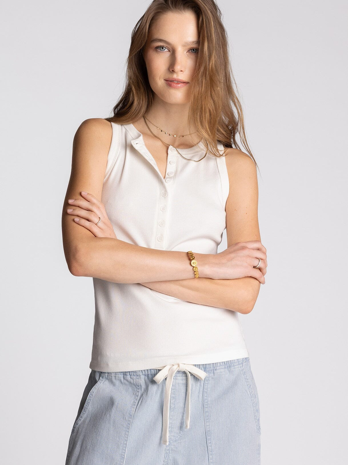 Woman wearing a white sleeveless top and light blue pants against a plain background