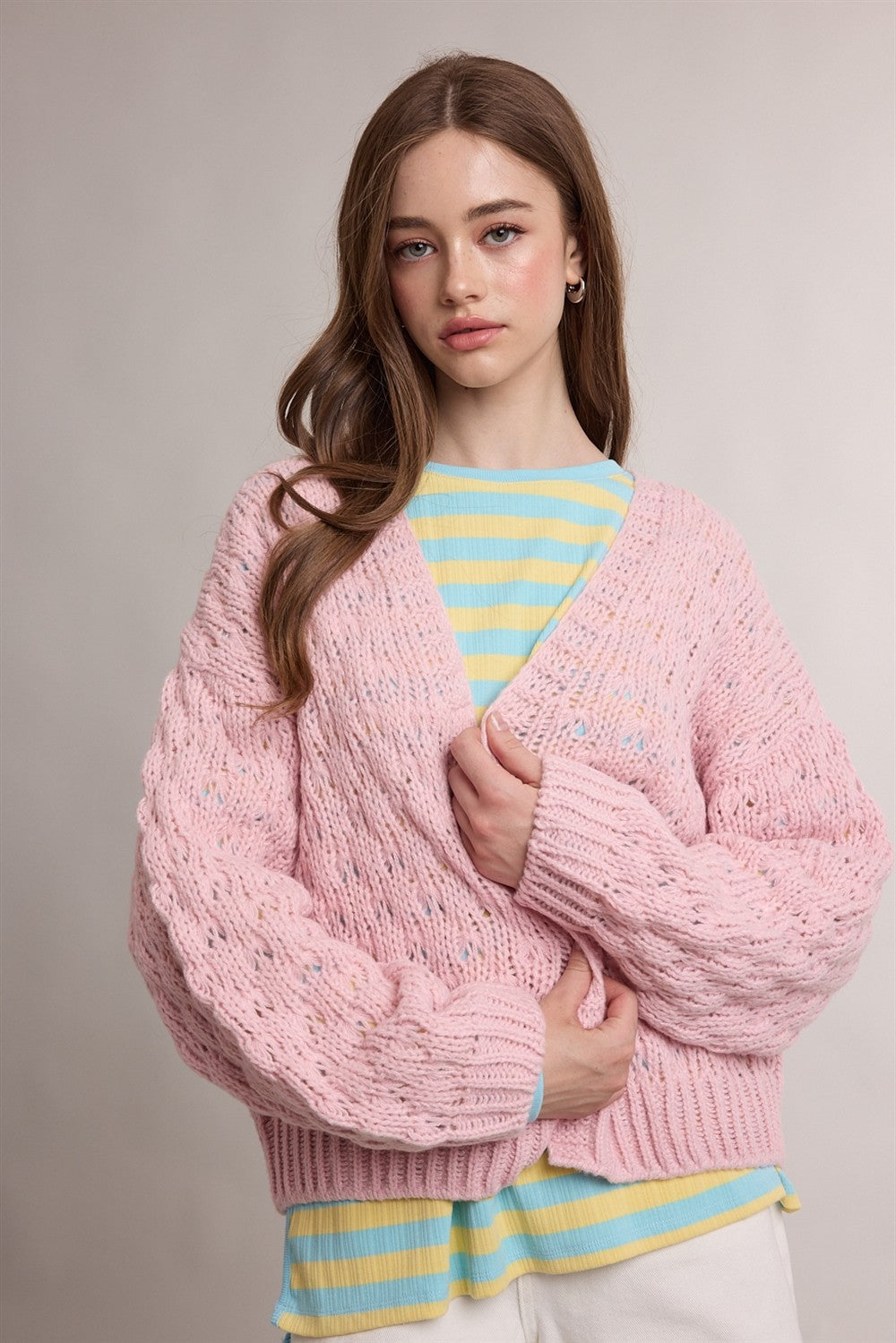A woman wearing a chunky pink cable-knit cardigan over a yellow and blue striped shirt, photographed against a neutral light gray studio background.