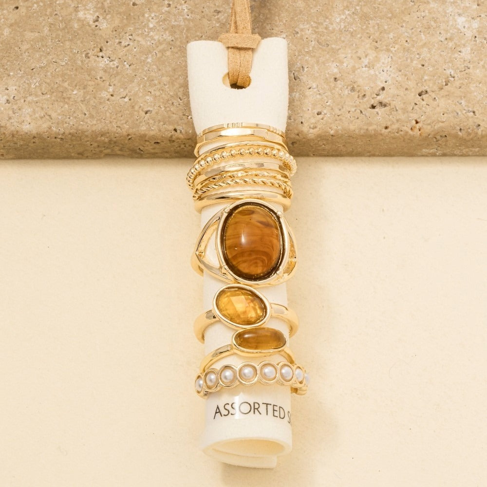 A stack of assorted gold rings displayed on a white ring holder, featuring various stone and metal designs, photographed against a neutral beige background.