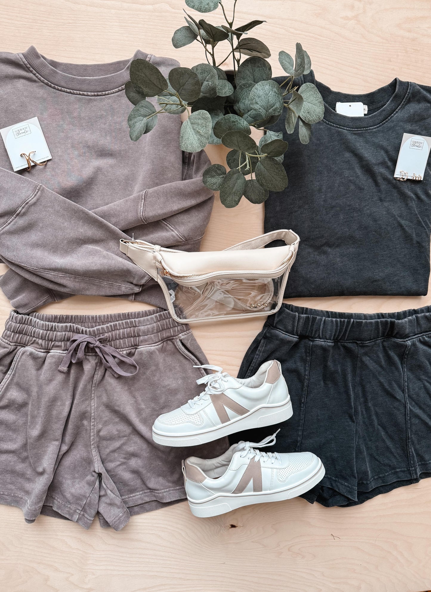 Two outfits laid out on a wooden surface with tags, a plant, and white sneakers.