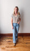 Woman wearing a striped shirt and jeans standing on a wooden floor with a white wall background