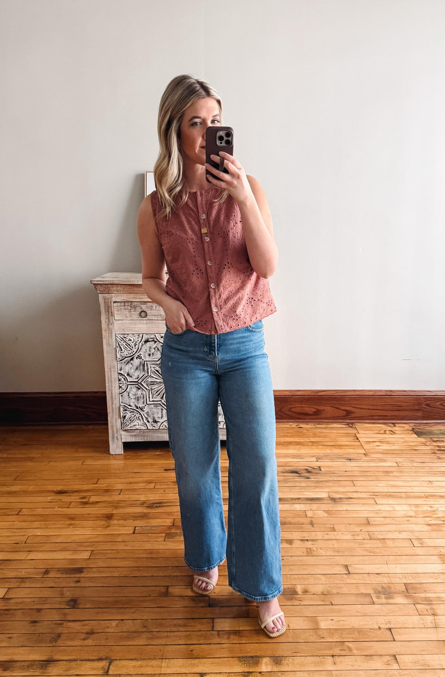 Woman taking a mirror selfie wearing a pink sleeveless shirt and blue jeans in a room with wooden flooring.