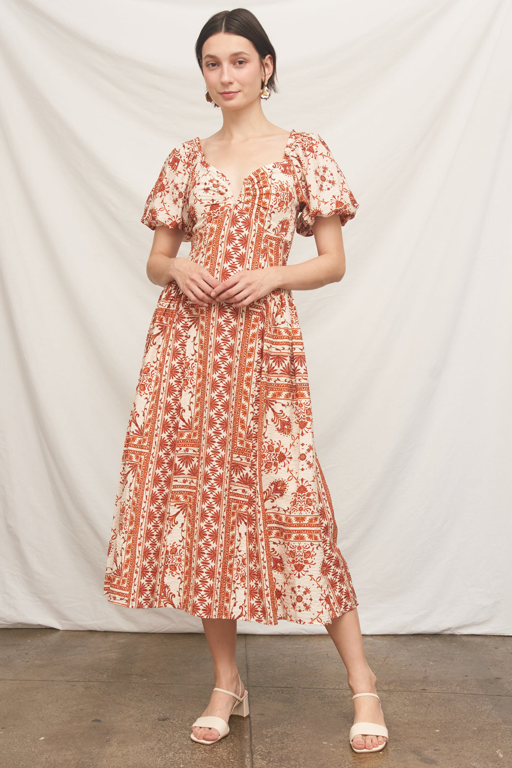 A woman modeling a short puff-sleeve midi dress with a rust-orange and cream floral/ornamental print, standing against a draped white fabric backdrop.