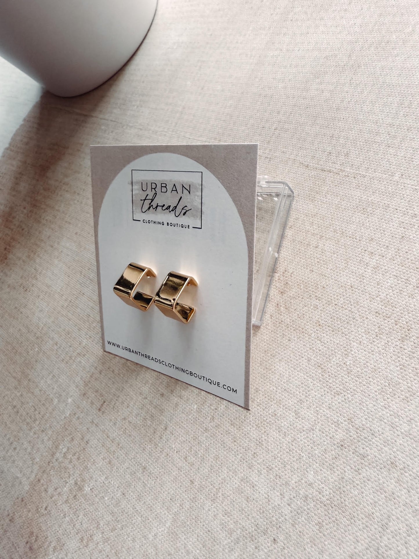 Gold earrings on a card with 'Urban Threads' branding on a beige surface.