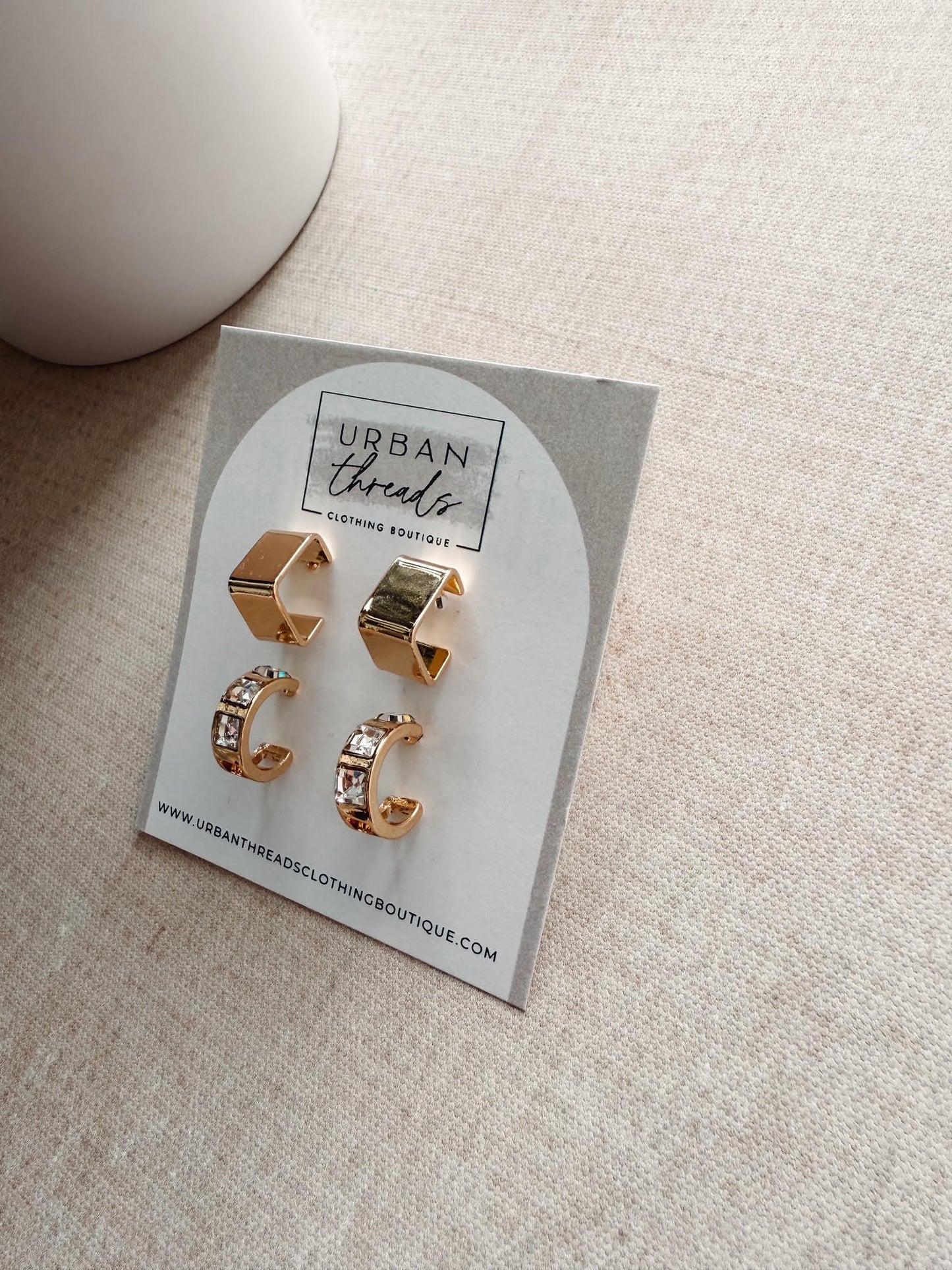 Gold earrings on a branded card with 'Urban Threads' on a beige surface