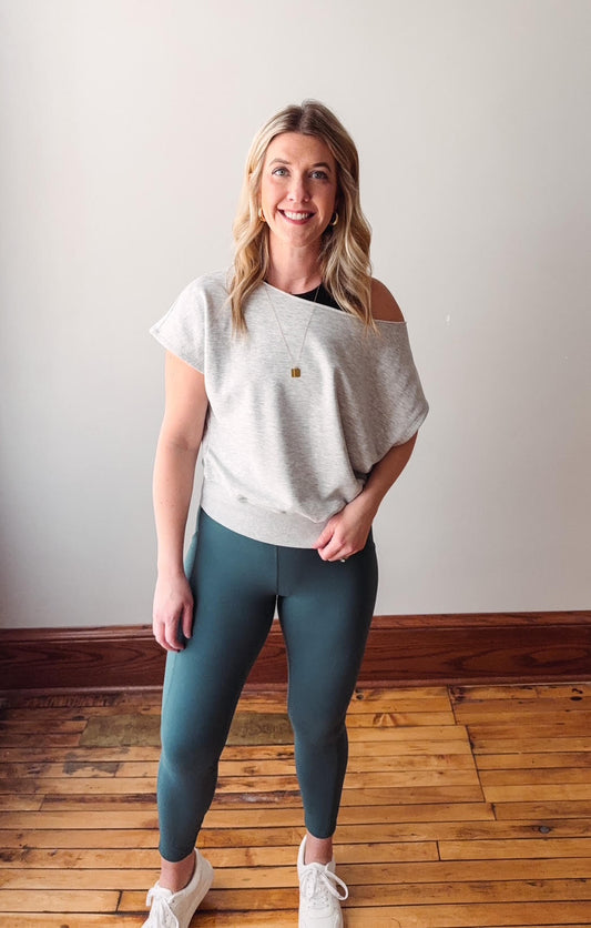 Woman wearing a light gray off-shoulder top and teal leggings standing on a wooden floor.