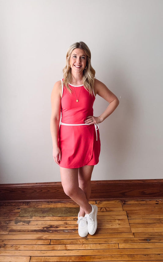 Woman wearing a red dress standing on a wooden floor against a plain wall.