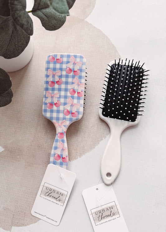 Two hairbrushes with decorative handles on a light surface, one with a floral pattern and the other with a black bristle design, branded 'Urban Threads'.