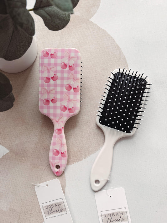 Two hairbrushes with decorative handles on a textured surface, one pink with cherry pattern and the other black bristle.