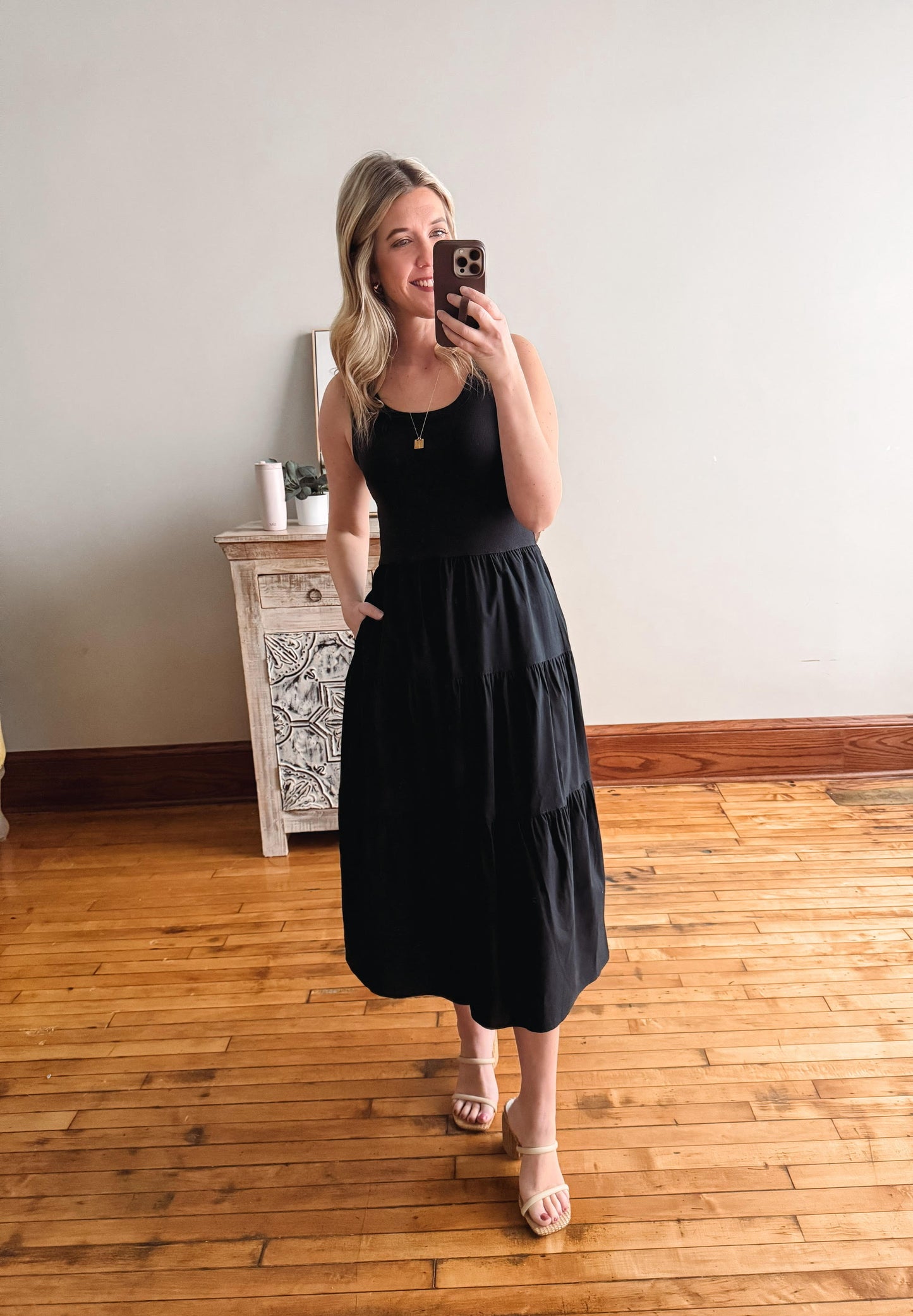 Woman taking a mirror selfie wearing a black dress in a room with wooden flooring.