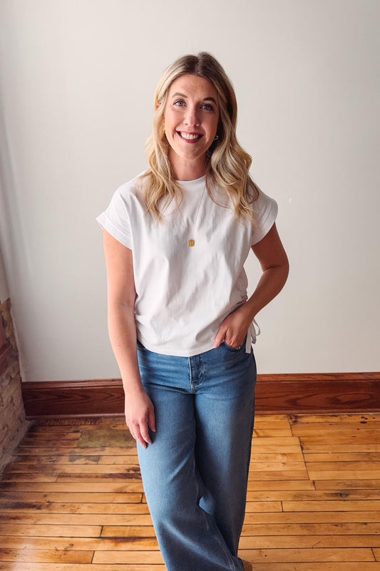 Woman wearing a white t-shirt and blue jeans standing in a room with wooden flooring and a white wall.