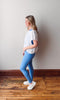 Woman wearing a white shirt and blue leggings standing against a plain wall.
