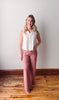 Woman wearing a white blouse and pink pants standing on a wooden floor with a white wall background