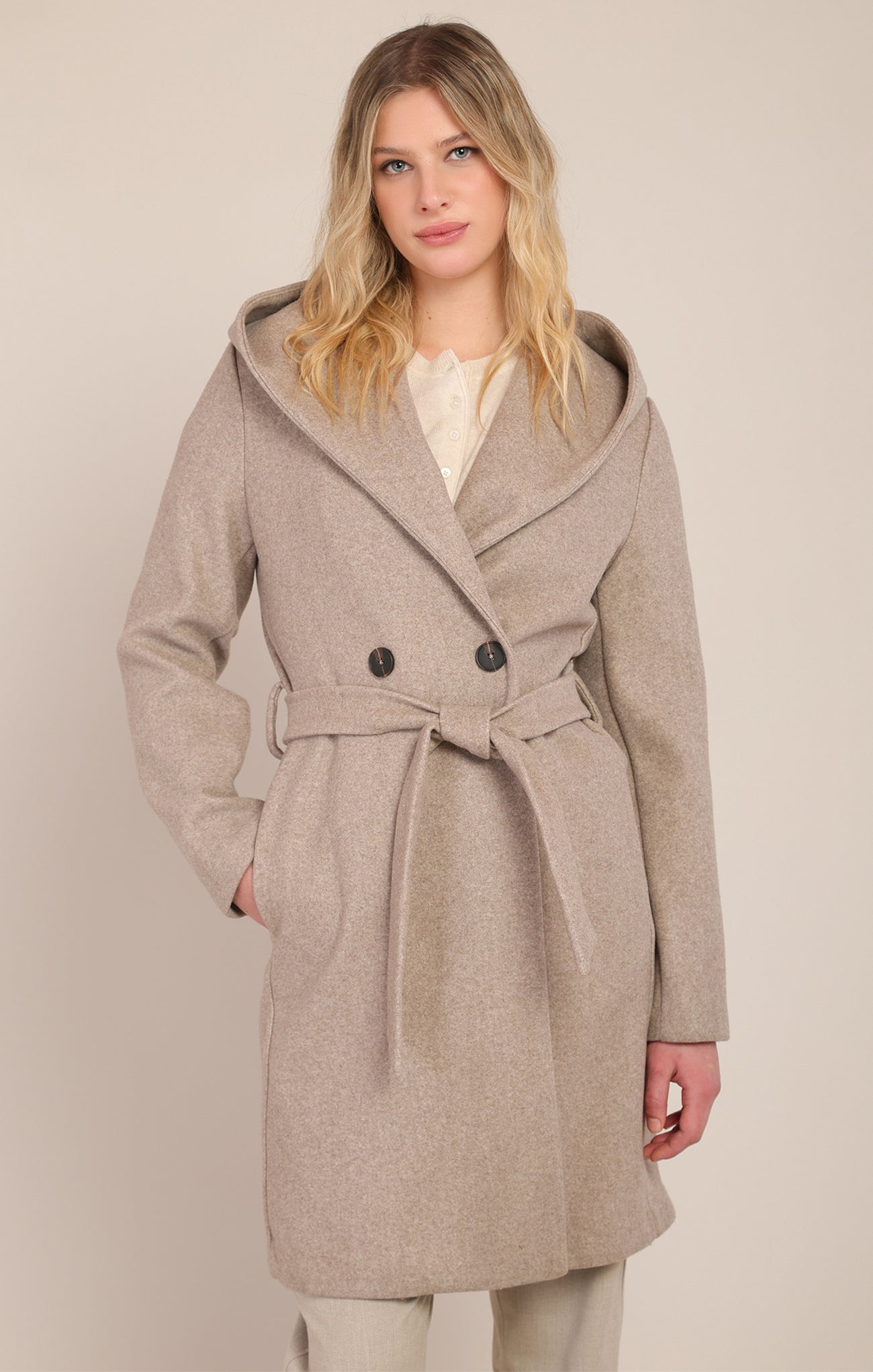 A woman wearing a light beige double-breasted hooded coat with a matching tie belt, styled over a cream top and light trousers, set against a plain beige studio background.