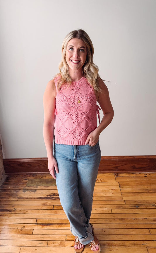 Woman wearing a pink sleeveless sweater and blue jeans standing on a wooden floor.