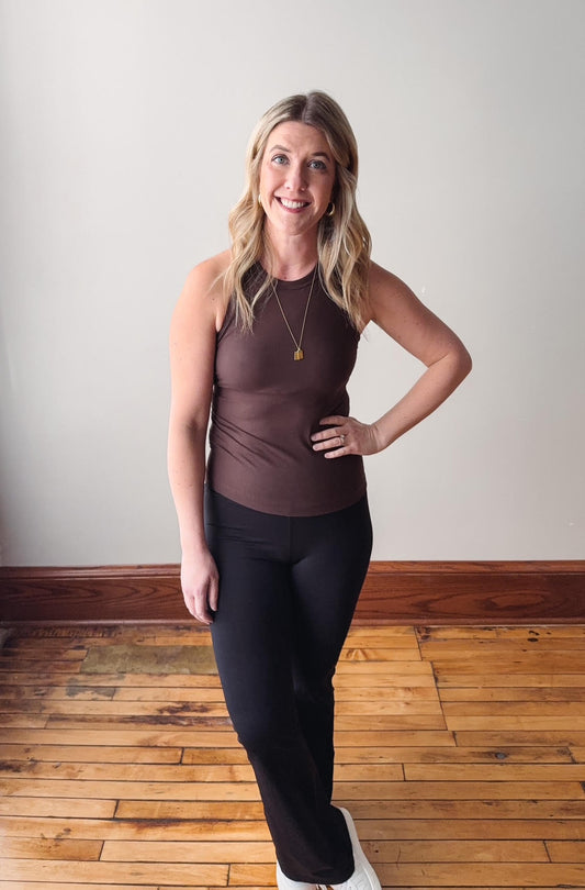 Woman wearing a brown tank top and black leggings standing on a wooden floor.