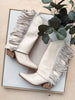 White cowboy boots with fringes on a light wooden surface with greenery.