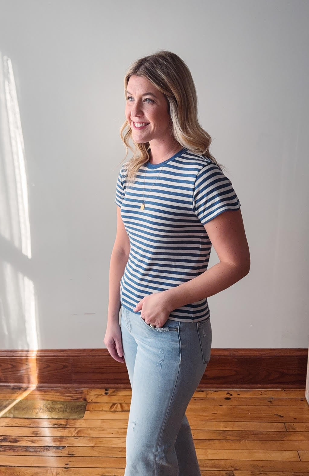 Woman wearing a striped shirt and jeans standing in a room with wooden flooring and a white wall.