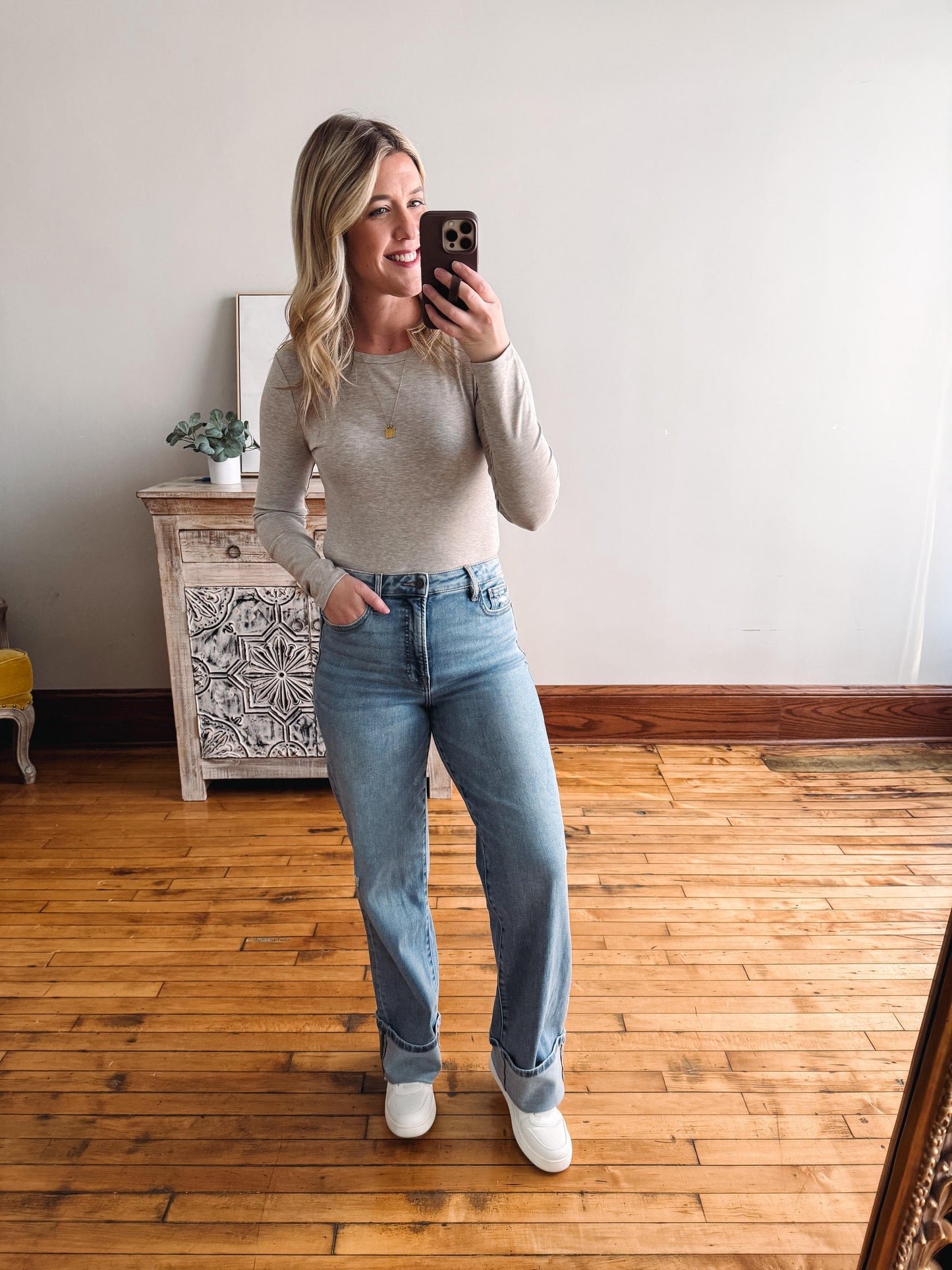 Woman taking a mirror selfie wearing a beige sweater and light blue jeans in a room with wooden flooring.