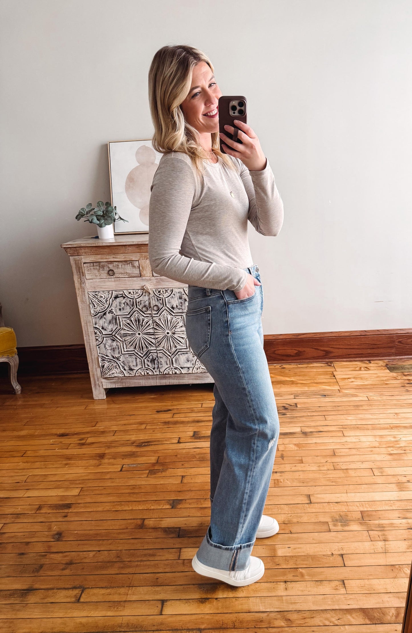 Woman taking a mirror selfie wearing a gray sweater and blue jeans in a room with wooden flooring and a decorative cabinet.