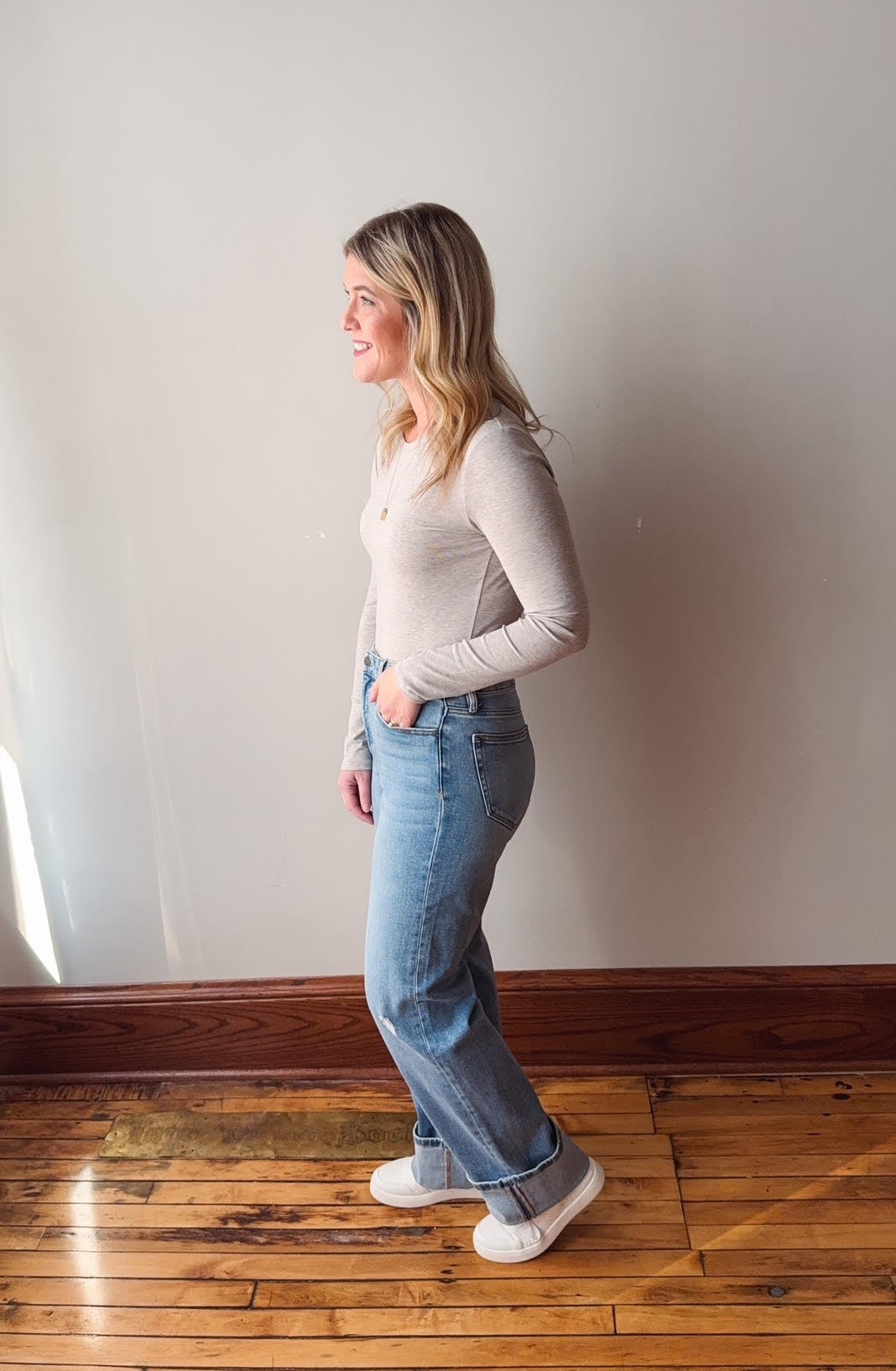Woman wearing a light gray long-sleeve top and blue jeans standing against a white wall.
