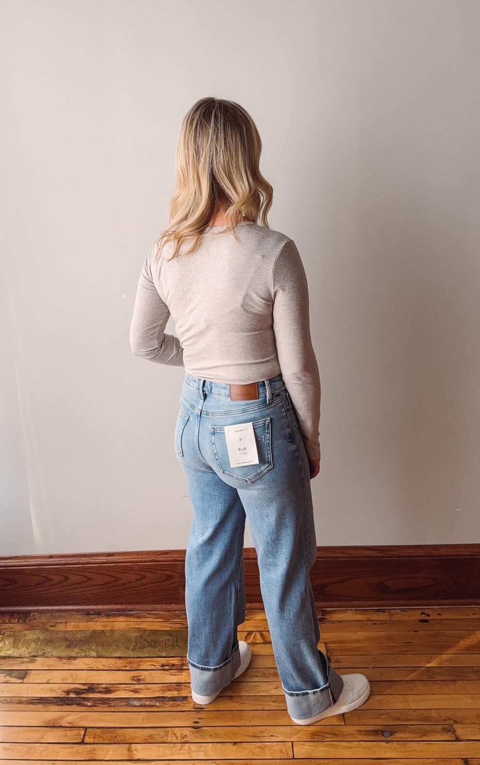 Person wearing light blue jeans and a beige sweater standing on a wooden floor with a plain wall background.