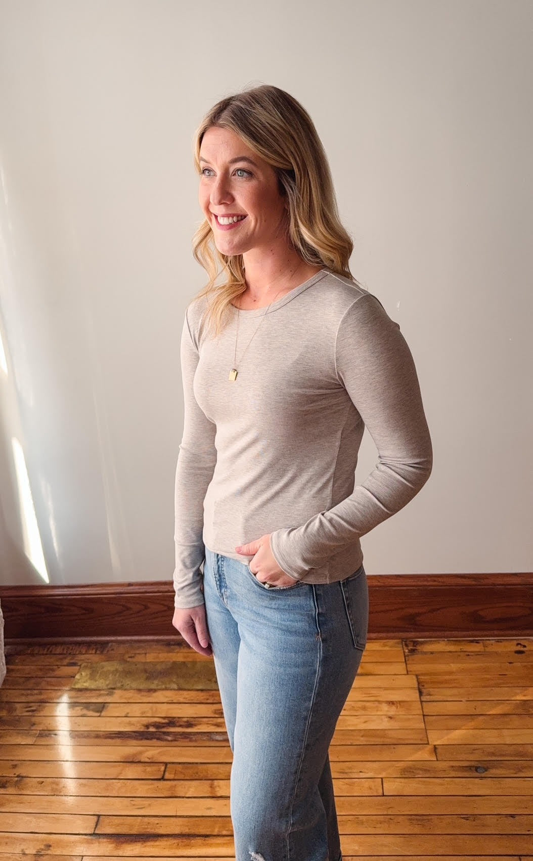Woman wearing a beige long-sleeve top and light blue jeans standing on a wooden floor.