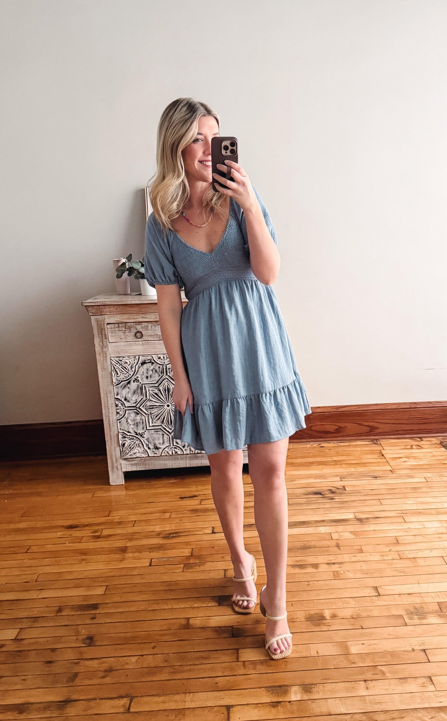 Woman taking a mirror selfie wearing a light blue dress in a room with wooden flooring and a decorative cabinet.