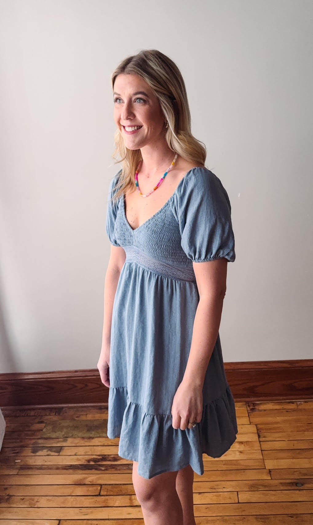Woman wearing a blue dress standing in a room with wooden flooring and a white wall.