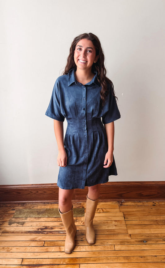 A woman wearing a short-sleeve button-front shirt dress in dark blue denim, paired with tan knee-high boots, standing on a wooden floor against a plain off-white wall.