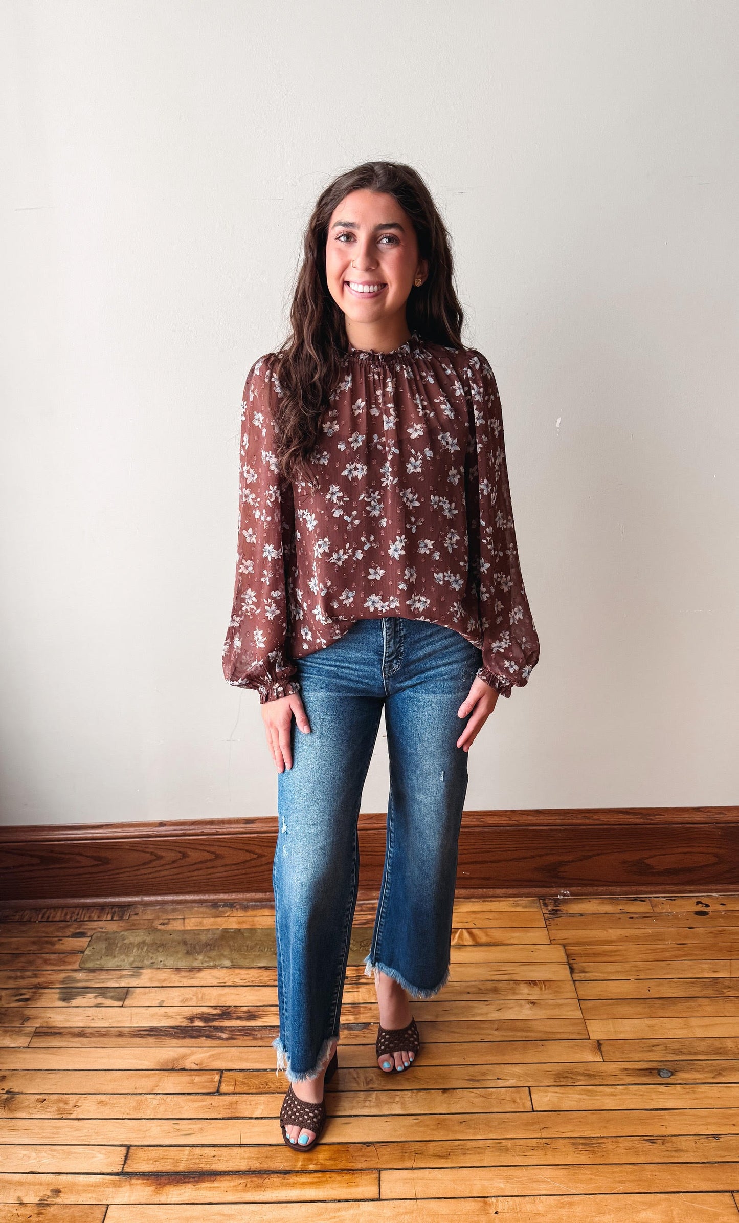 A woman stands in a room with a light beige wall and wooden floor, wearing a brown long-sleeve blouse with a white floral pattern, blue jeans, and brown patterned sandals.