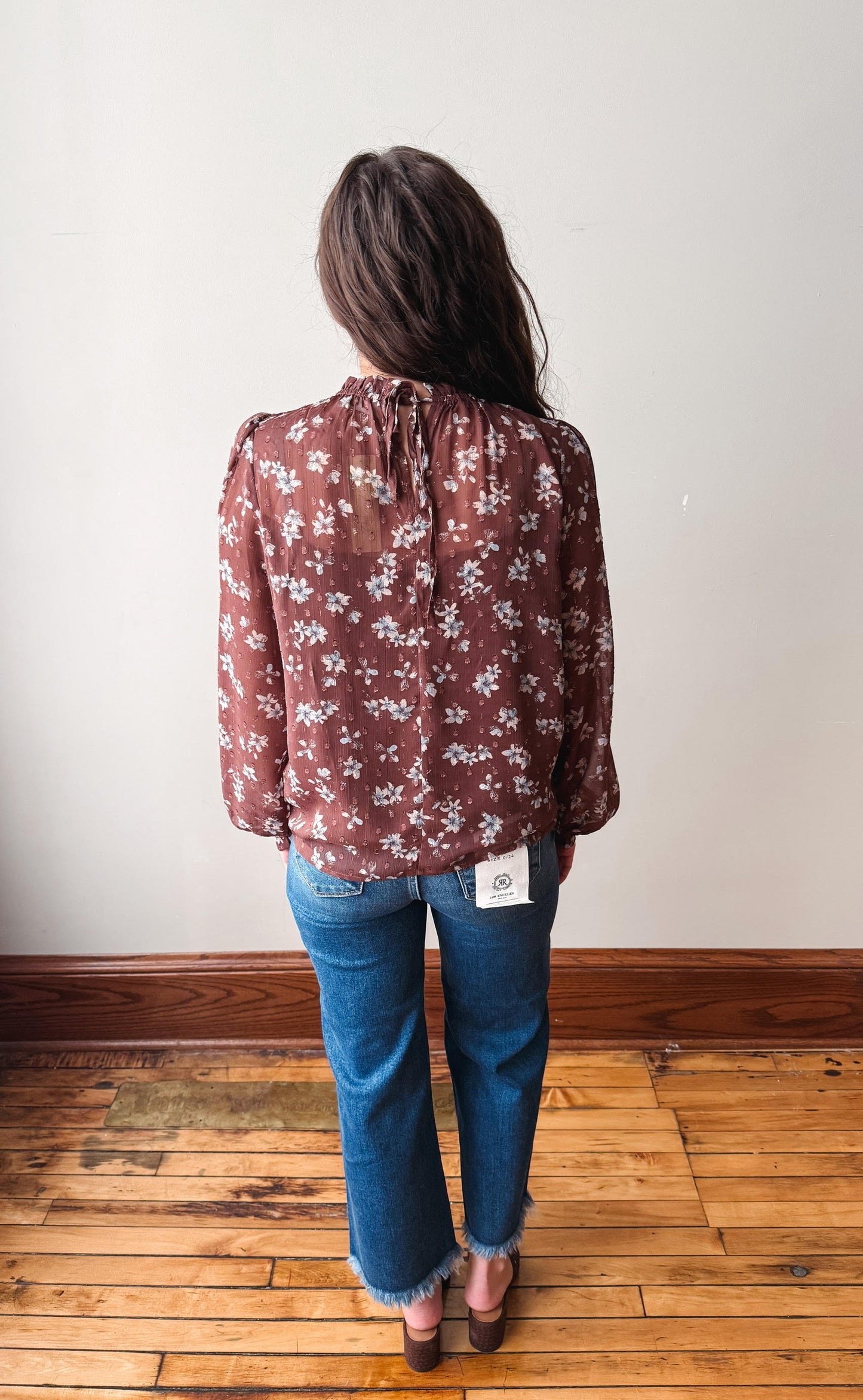 A woman viewed from behind wearing a rust-brown long-sleeve blouse with a white floral pattern, paired with blue jeans and brown shoes, standing on a wooden floor against a plain off-white wall.