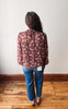 A woman viewed from behind wearing a rust-brown long-sleeve blouse with a white floral pattern, paired with blue jeans and brown shoes, standing on a wooden floor against a plain off-white wall.
