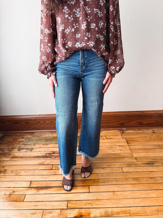 A person wearing blue straight-leg jeans with a raw frayed hem, paired with a brown floral blouse and brown strappy heels, standing on a wooden floor against a plain wall.