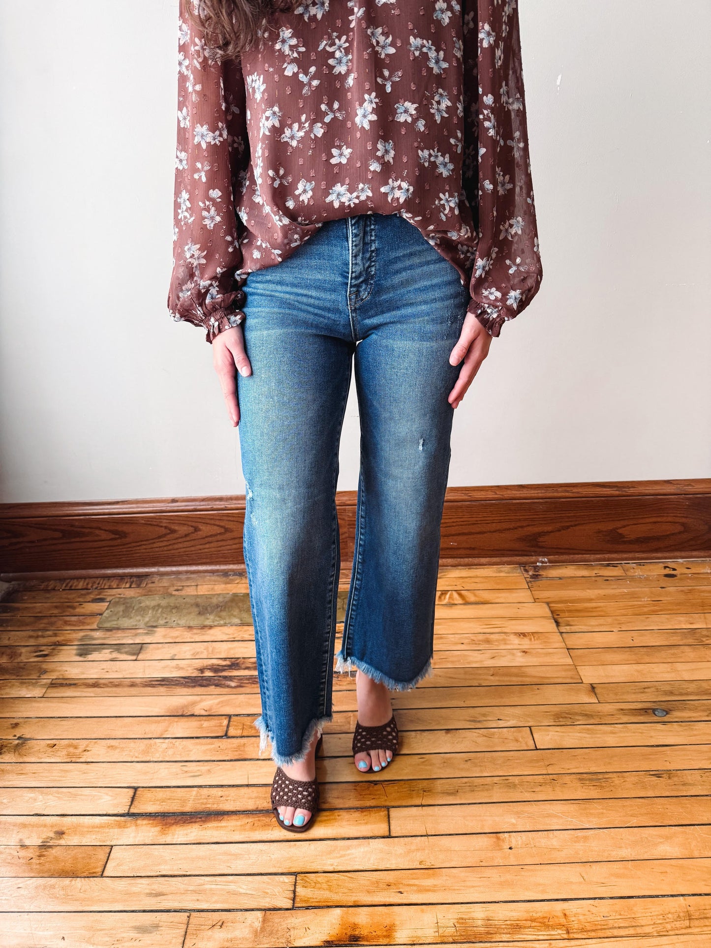 A person wearing blue straight-leg jeans with a raw frayed hem, paired with a brown floral blouse and brown strappy heels, standing on a wooden floor against a plain wall.