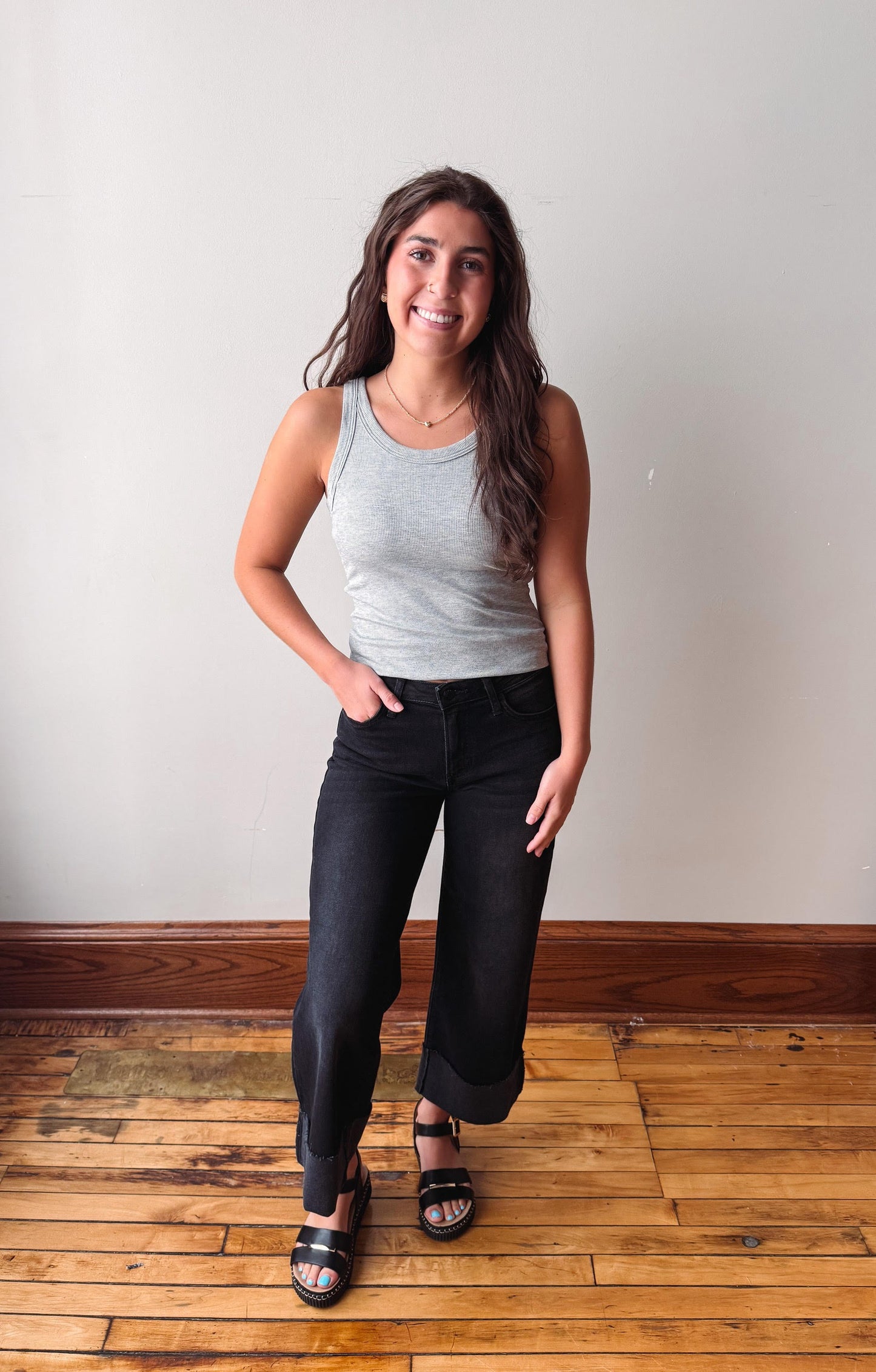 A woman modeling a light gray ribbed tank top and black wide-leg jeans with black sandals, standing on a wooden floor against a plain off-white wall.