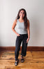 A woman modeling a light gray ribbed tank top and black wide-leg jeans with black sandals, standing on a wooden floor against a plain off-white wall.